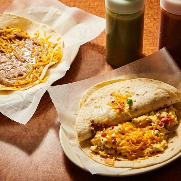 Two breakfast tacos, one with beans and cheese, the other with scrambled eggs, on a wooden table.