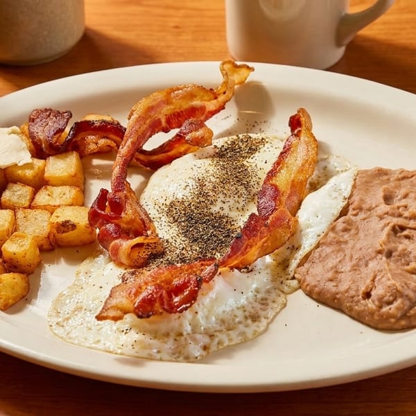 Breakfast plate with fried eggs, crispy bacon, home fries, refried beans, and tortillas on a wooden table.