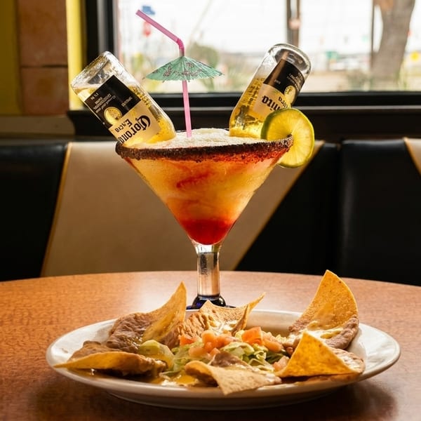 A large margarita with two inverted Corona bottles and a plate of nachos on a wooden table.