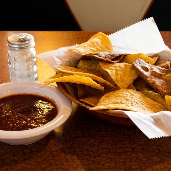 A basket of tortilla chips with a bowl of salsa and a salt shaker on a wooden table.