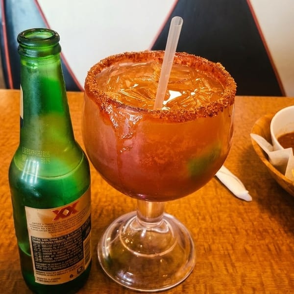 A Michelada, beer bottle, and a basket of chips with salsa on a wooden table.